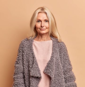Fashionable fifty years old woman with blonde hair dressed in jumper and warm coat looks directly at camera with serious expression poses against beige background stays beautiful in any age.